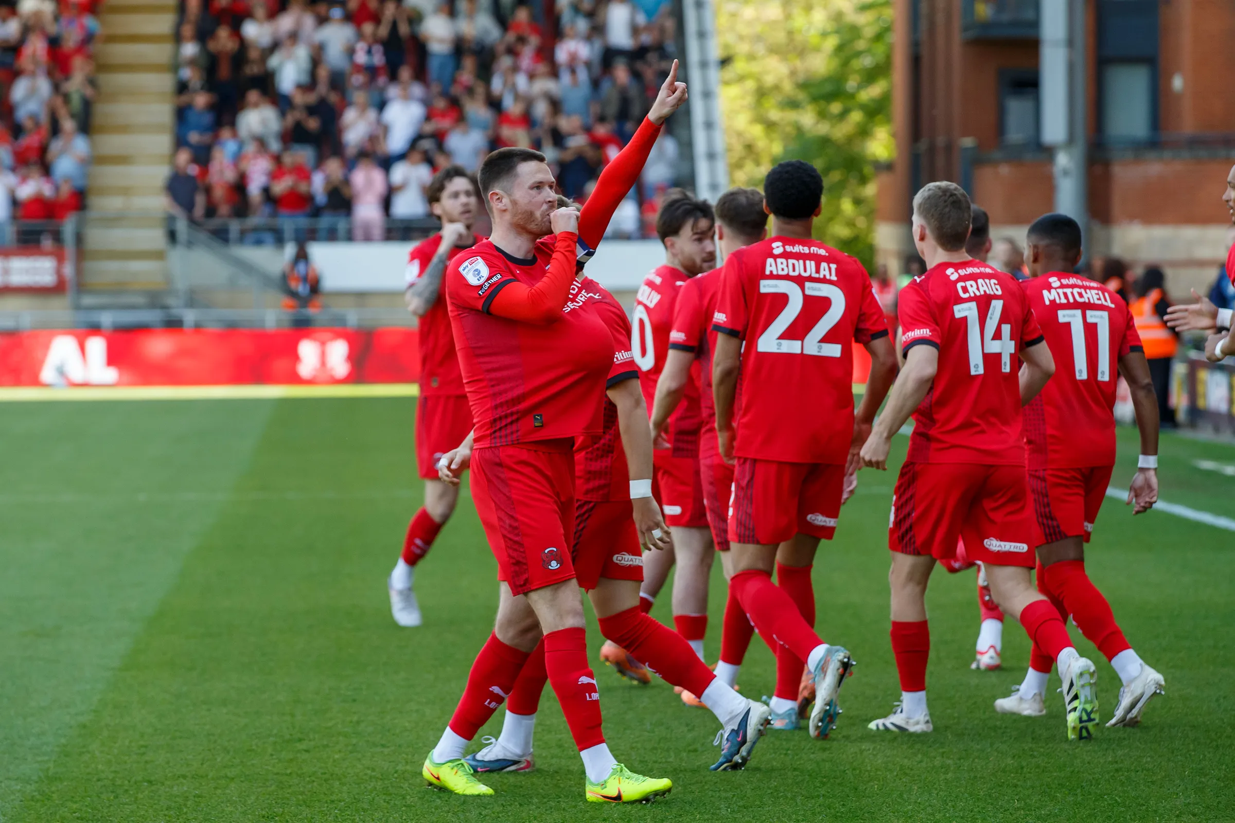 Leyton Orient players in red celebrating on the pitch as supporters watch from the stand at Brisbane Road