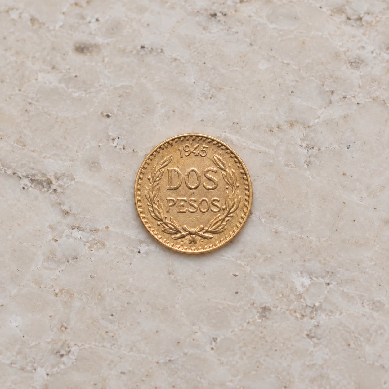 A complete small 1945 Mexican 2 Pesos gold coin with a wreath design on pale limestone