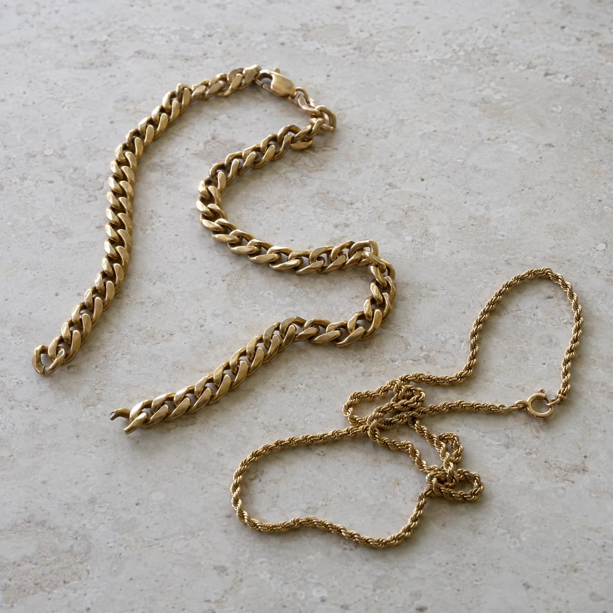A broken gold curb chain and tangled fine rope chain on pale stone