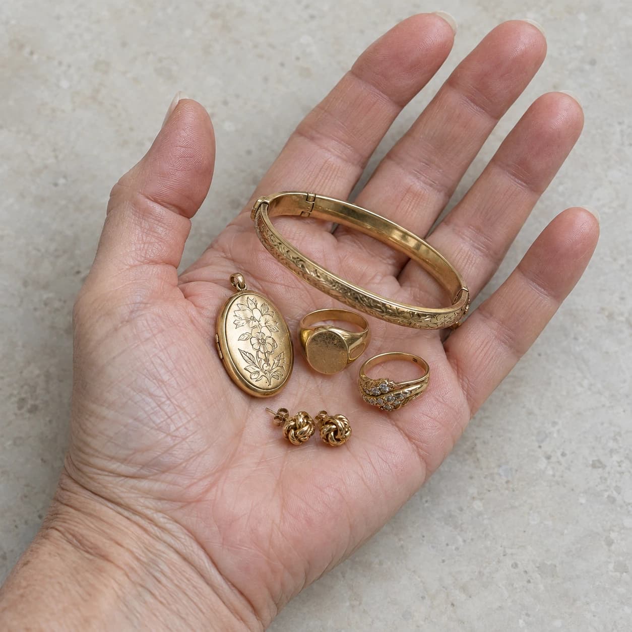 An open hand holding a gold bangle, locket, rings and stud earrings